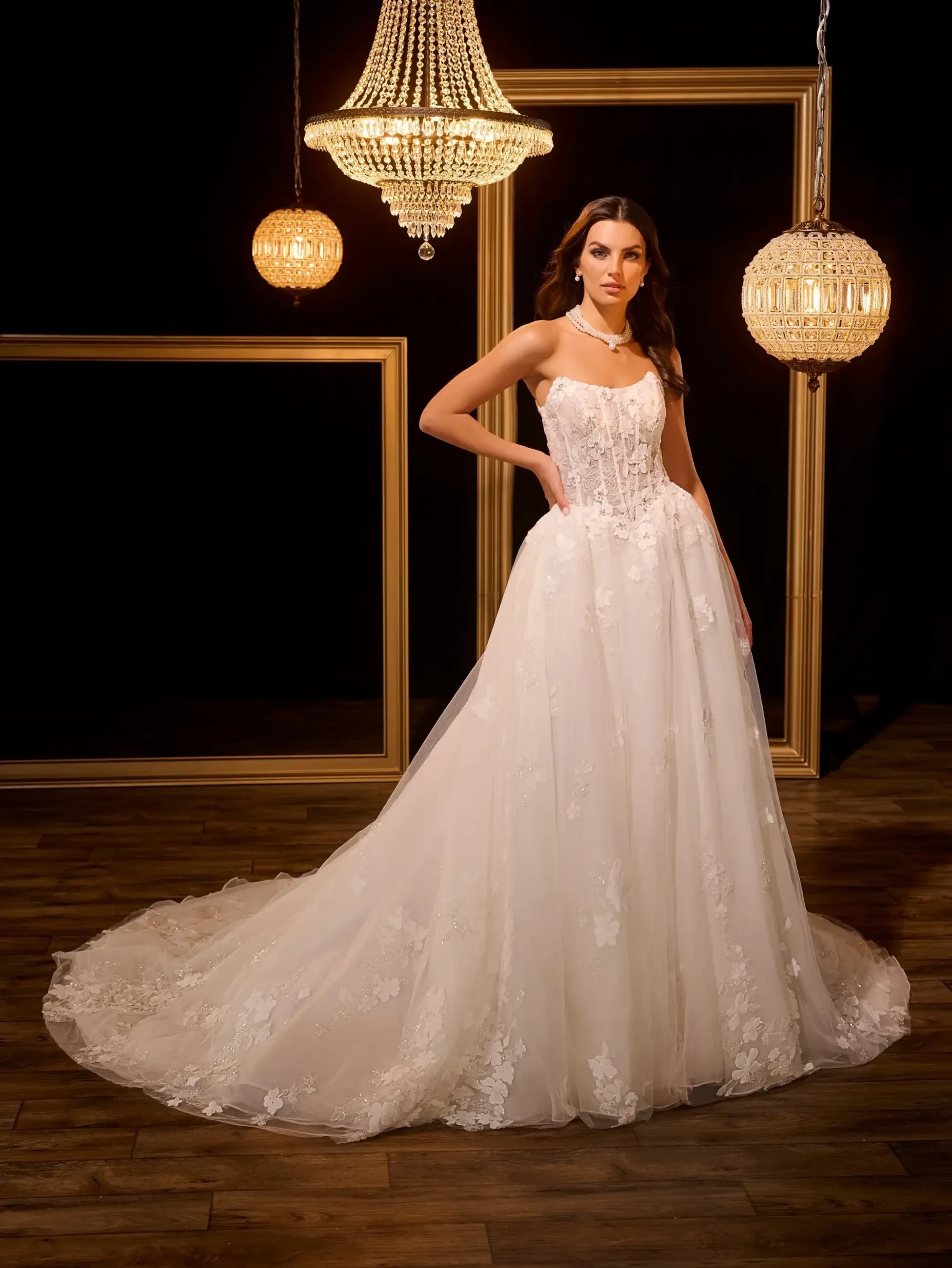 A model wearing a stunning white wedding gown with intricate lace details, standing elegantly in a dimly lit room adorned with vintage chandeliers and golden picture frames.