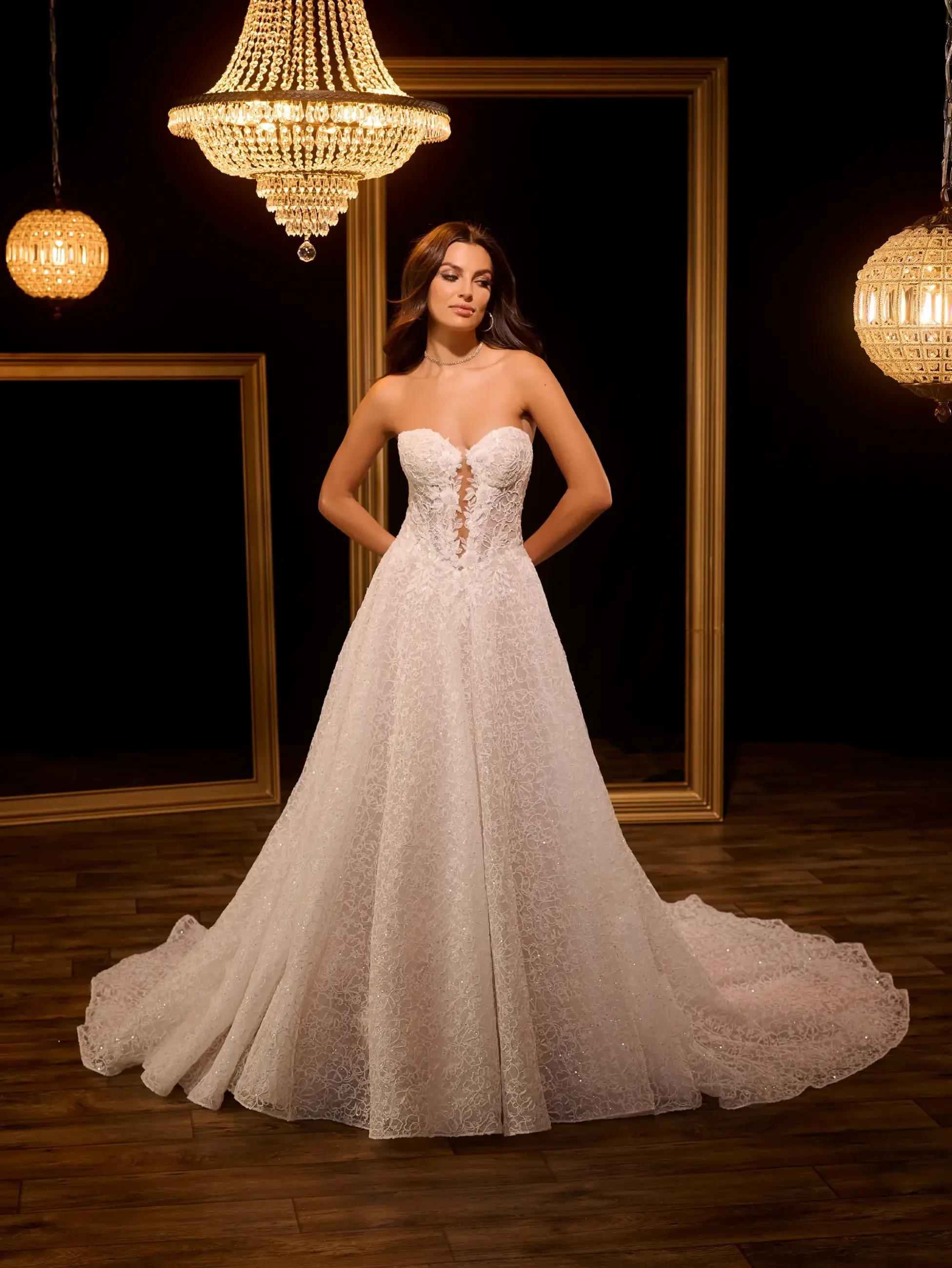 Elegant woman in a strapless lace wedding gown stands confidently on a warm, lit stage with chandeliers and wooden floor, exuding luxury and grace.