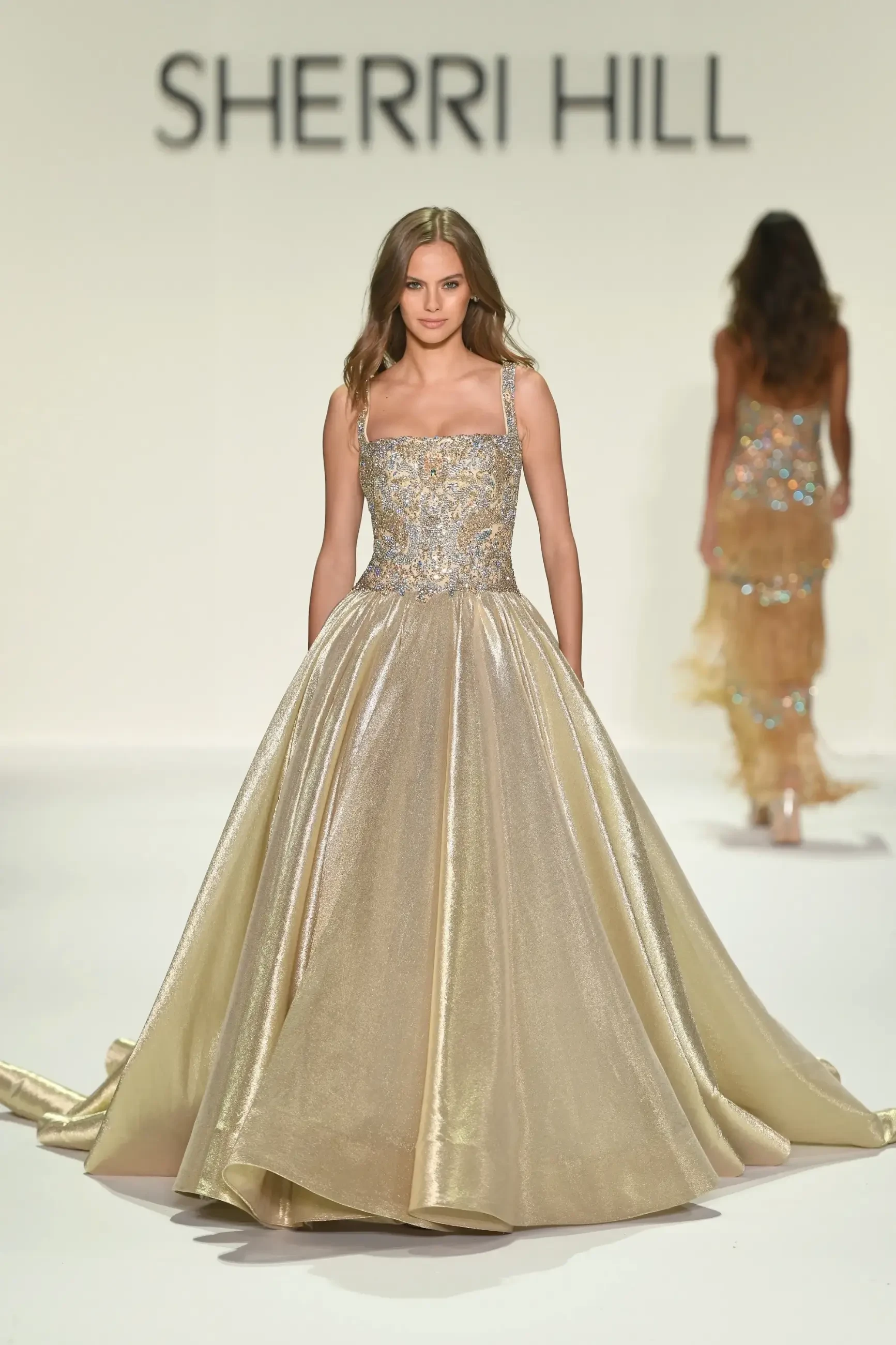 A model walks a runway in a shimmering gold gown with a fitted, sequined bodice and full skirt. The background displays 