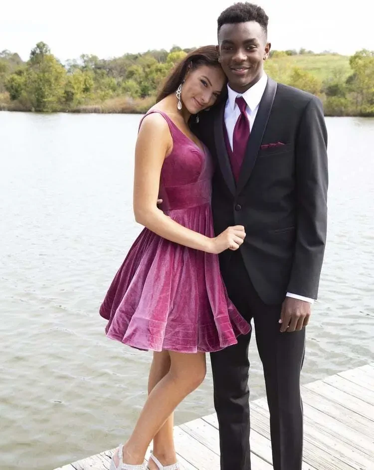 Two young individuals dressed formally stand close together on a dock by a body of water. The person on the left wears a sleeveless, purple velvet dress, while the individual on the right is in a black suit with a maroon tie. Green trees and hills are visible in the background.