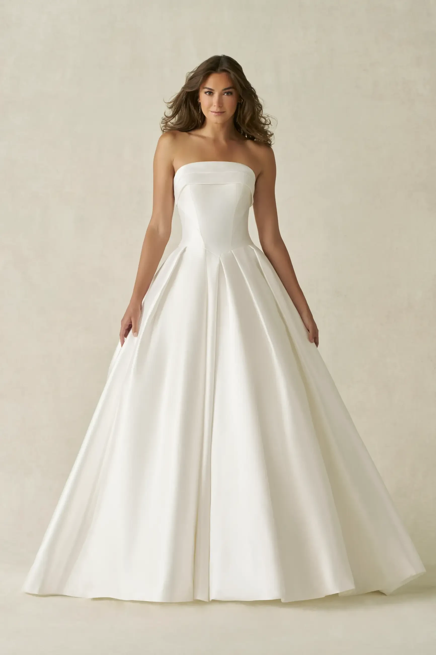A woman stands wearing a strapless, A-line satin wedding gown with a fitted bodice and flowing skirt. The background is plain, highlighting the elegant dress.