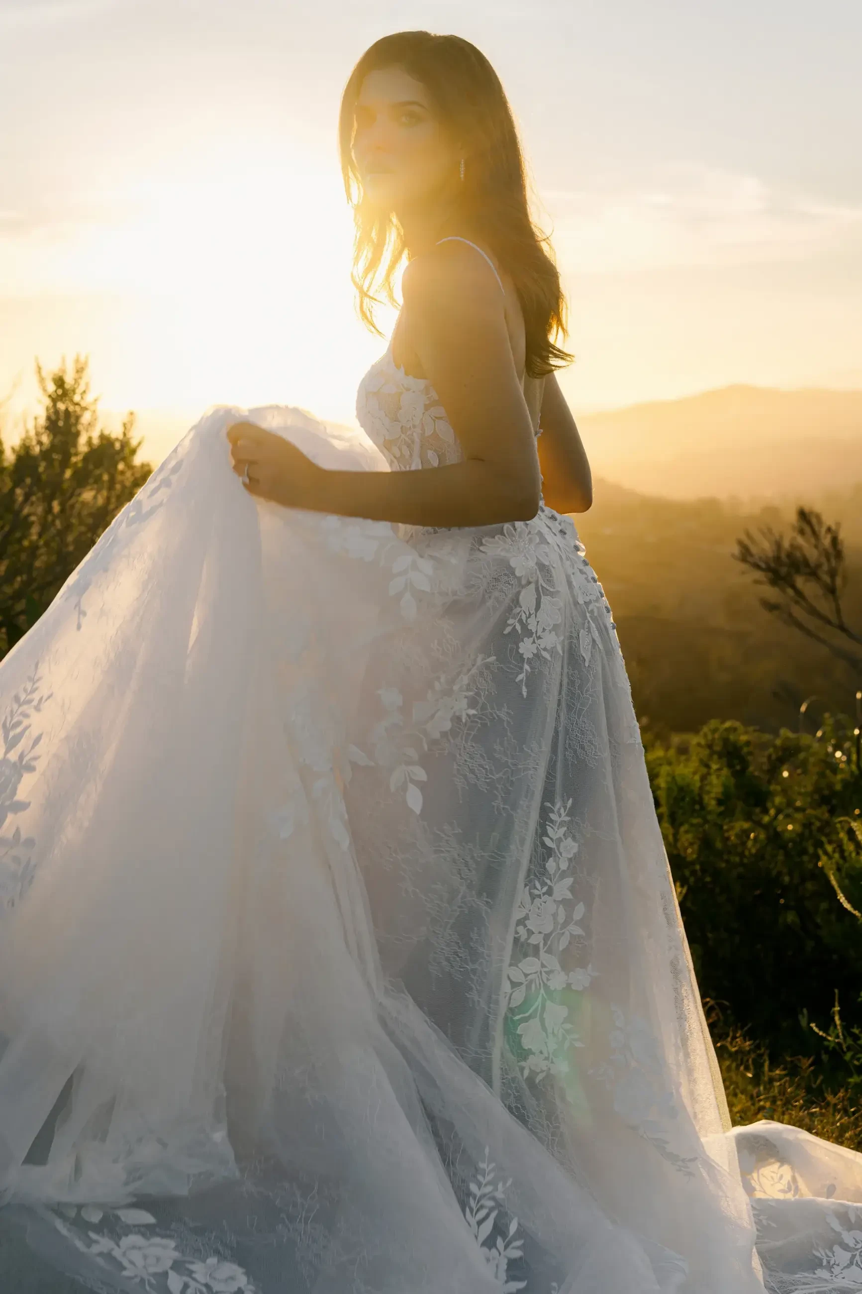 A woman in a flowing lace wedding dress stands outdoors at sunset. The sunlight creates a warm glow, highlighting the dress's intricate floral patterns.