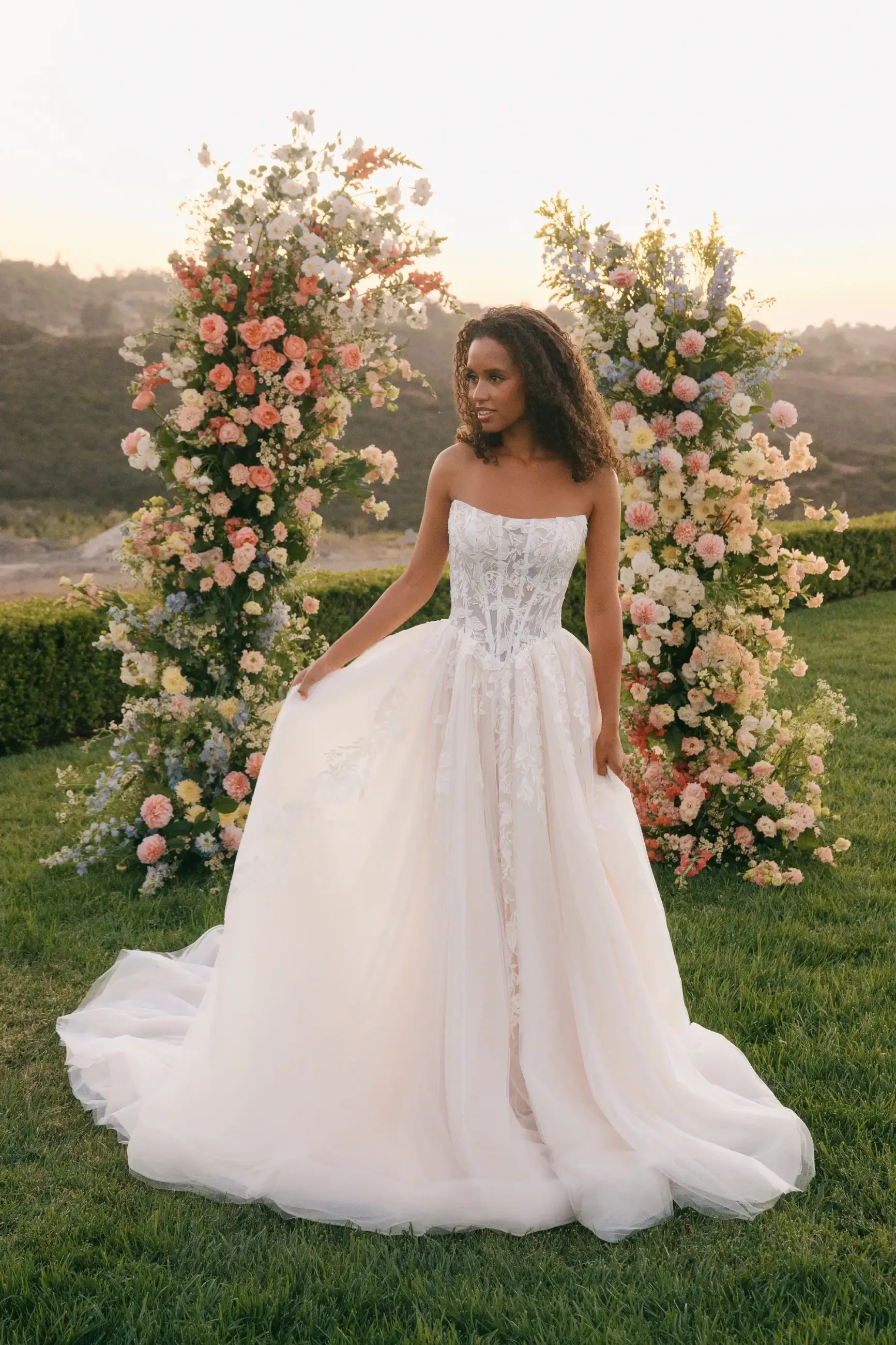 A bride in an elegant, strapless white gown stands on grass, framed by tall, colorful floral arrangements. The background shows rolling hills at sunset.