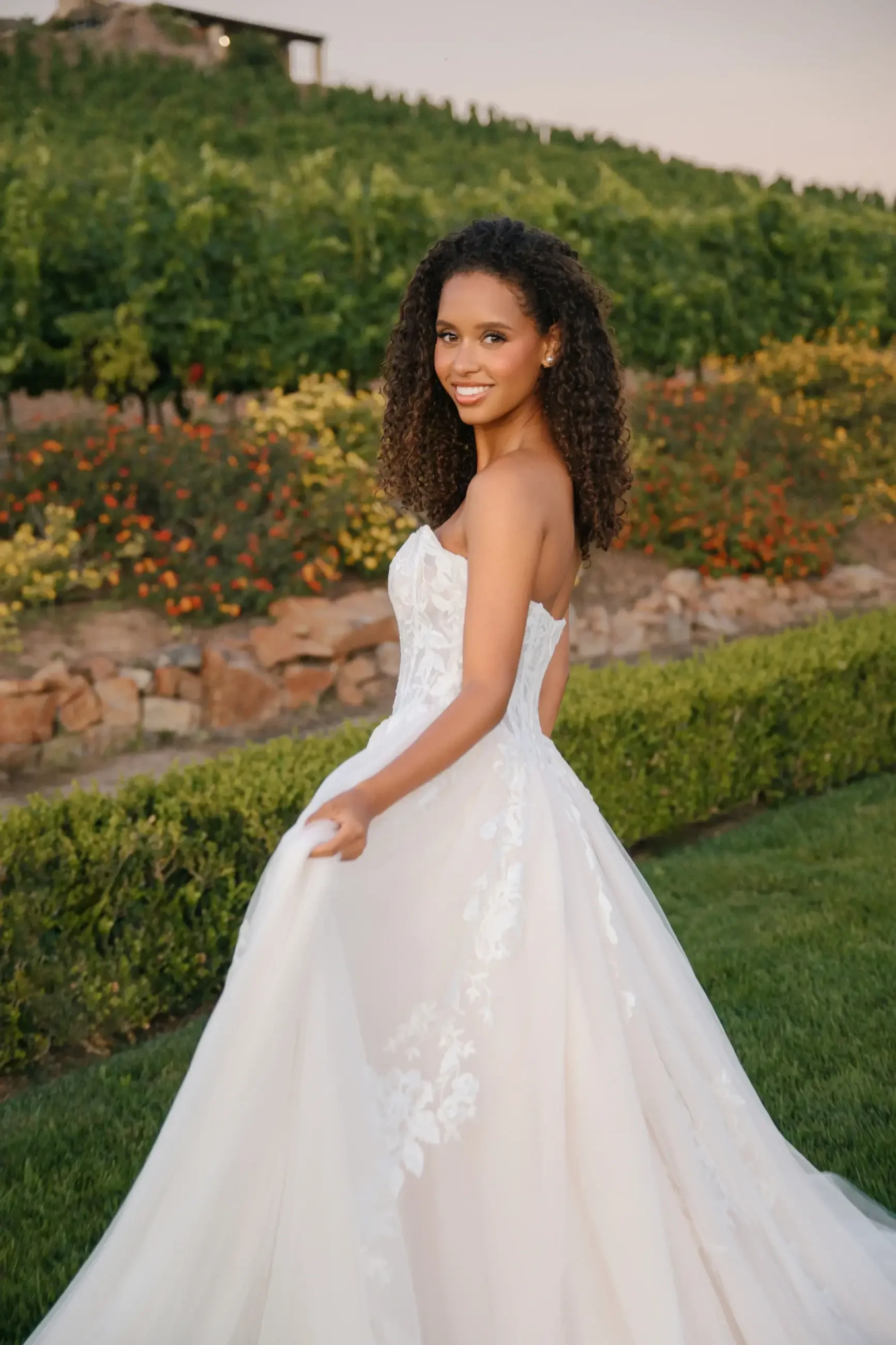 Bride in a flowing white gown smiles radiantly, standing in a vibrant garden with lush greenery and colorful flowers, creating a joyful atmosphere.