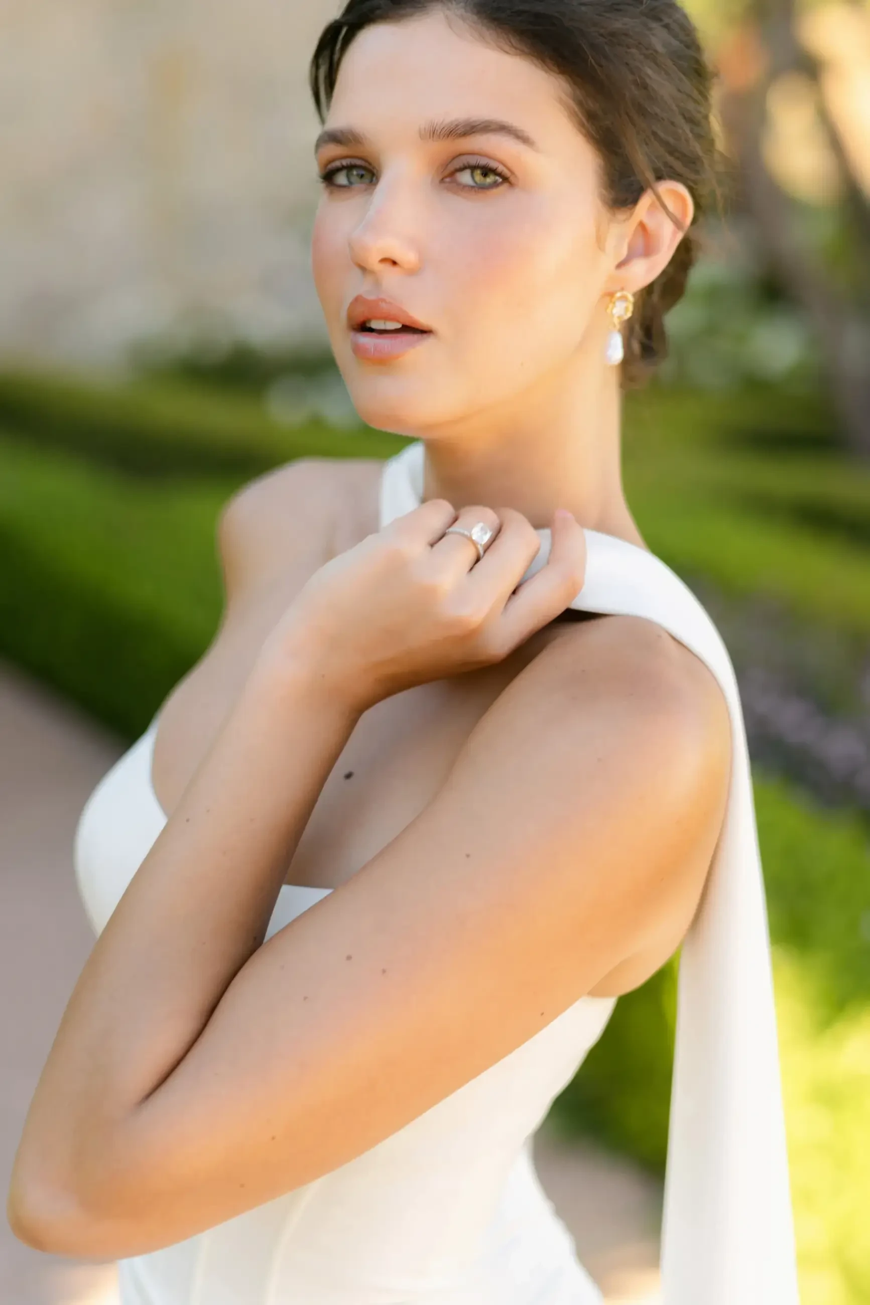 A woman in a white dress with a strap and a flowing fabric draped over her arm, posing outdoors with greenery in the background.
