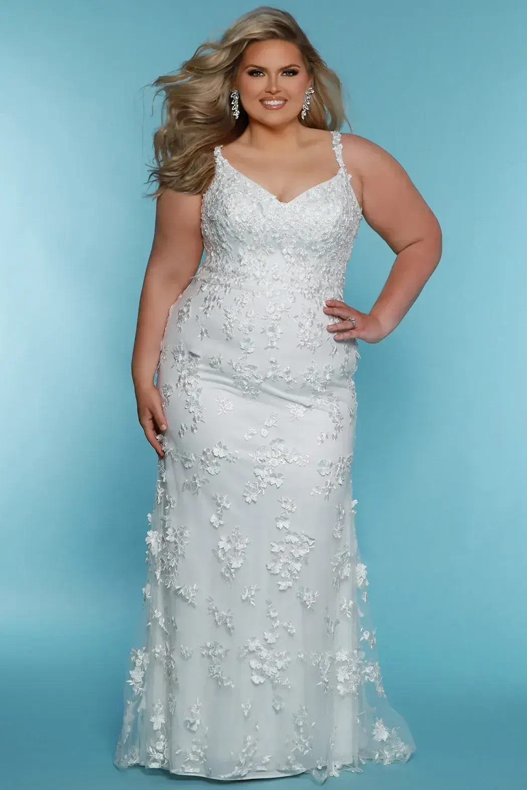 A woman in a white, floral-embellished gown stands against a light blue background, smiling and posing with one hand on her hip. She has long hair and wears earrings.