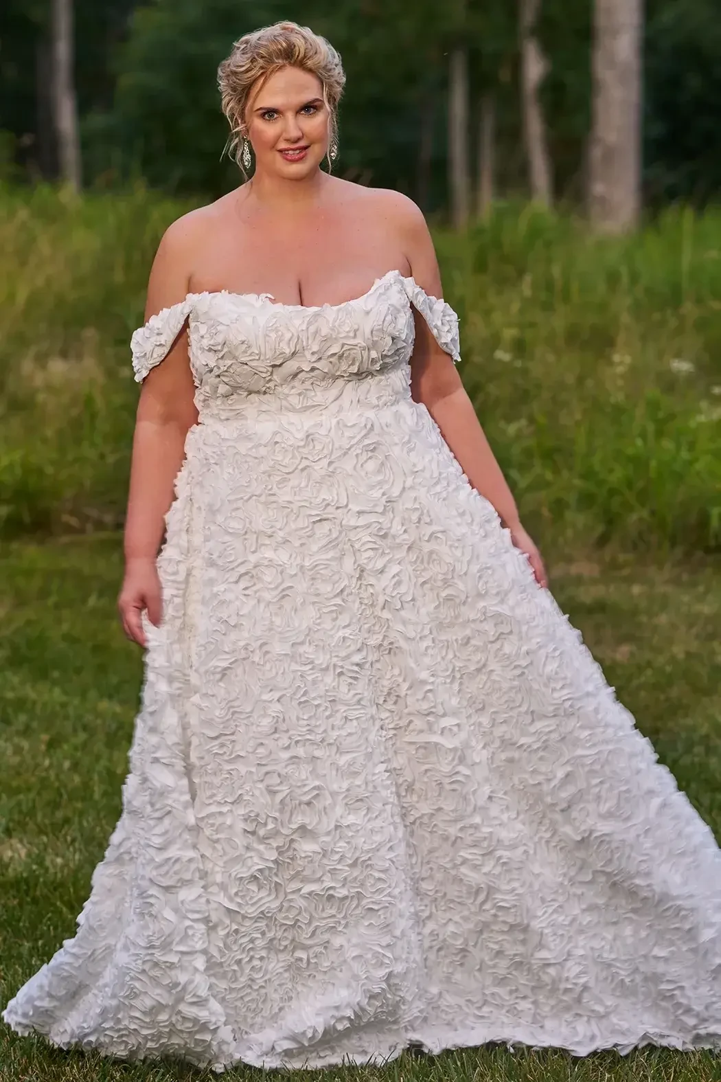 A woman wearing an off-the-shoulder wedding dress with floral embellishments, standing in a grassy area with trees in the background.