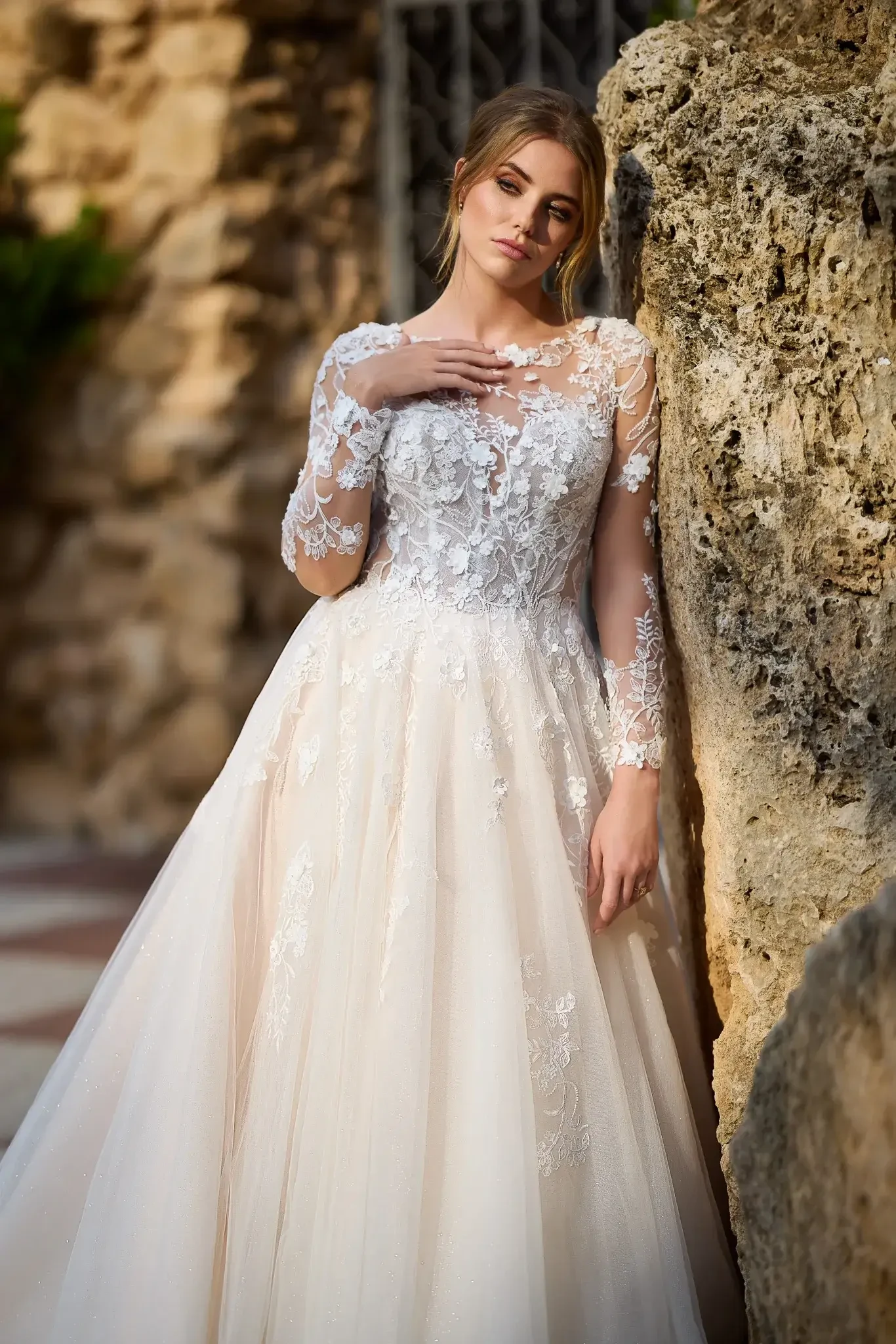 Bride in lace gown with floral patterns leans against a textured stone wall. She gazes softly, evoking a serene and romantic atmosphere.